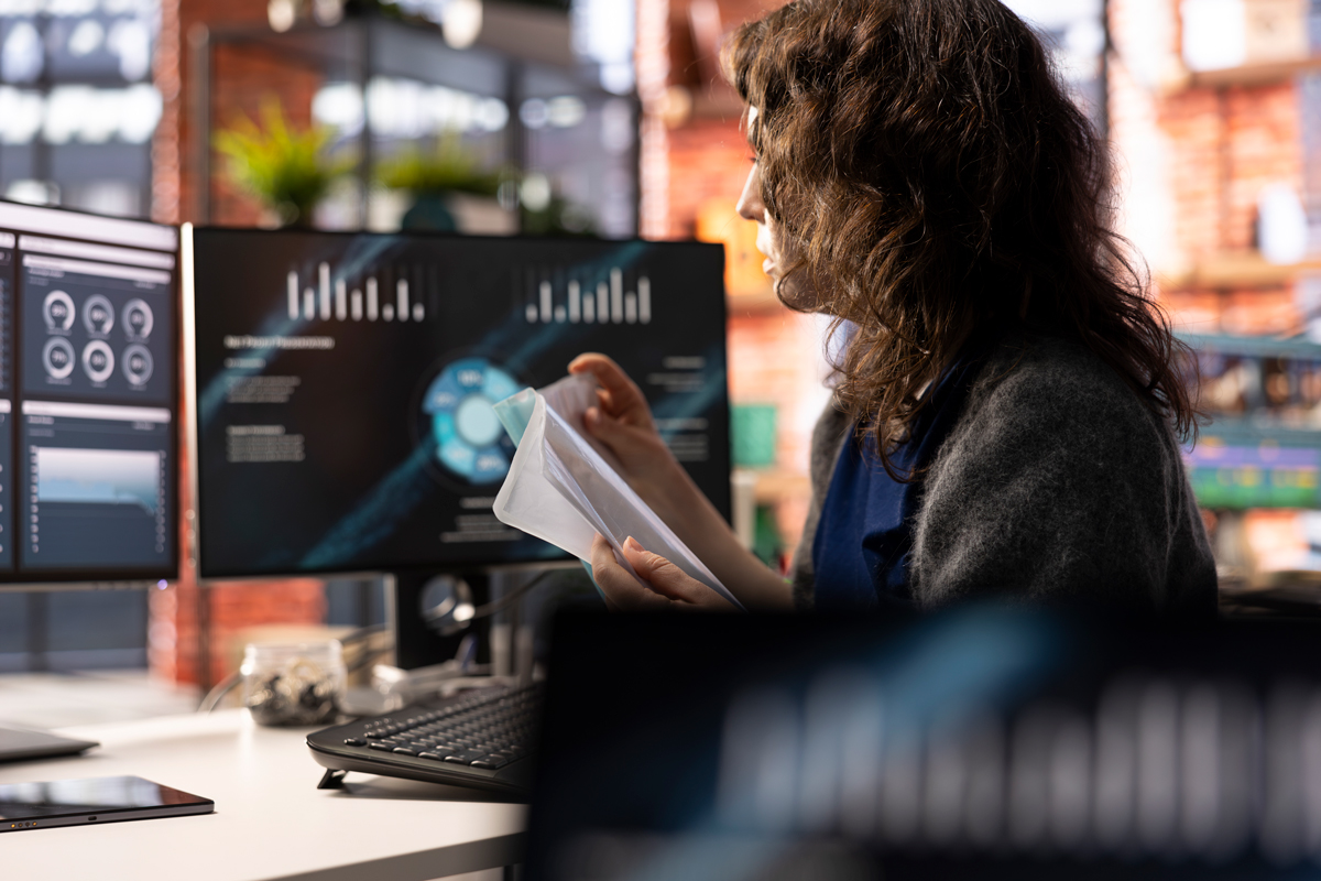 Woman with curly dark hair reviewing printed documents at a multi-monitor analytics workstation